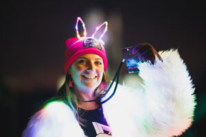 A person holds up a volunteer lanyard at ILLUMINATE: Light Art and Technology Festival. She's wearing a light up jacket and a bright pink ILLUMINATE beanie.